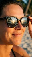 Middle-aged Caucasian male wearing dark-framed sunglasses with tropical beach sunset reflection in lenses, smiling at camera on sandy shore during golden hour.