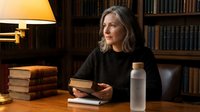 Middle-aged Caucasian woman reading book in cozy library setting with warm lamp lighting and bookshelf background for educational content.