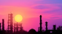 Industrial oil refinery complex silhouetted against vibrant pink and purple sunset sky with bright yellow sun for energy and petroleum industry concepts.