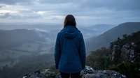 Woman with long dark hair wearing a blue jacket standing on rocky mountain peak overlooking misty valley landscape, rear view, outdoor adventure and hiking concept.