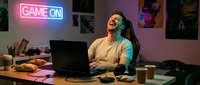 Young woman with long dark hair, wearing headphones and casual clothing, laughing while gaming at laptop desk with neon game on sign, snacks and coffee mug nearby.