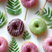 Colorful glazed donuts with drizzle decoration and tropical palm leaves arranged on white background, creating modern food composition for bakery marketing.