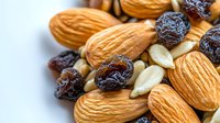 Fresh raw almonds mixed with dark raisins and peeled pumpkin seeds on white background, macro food photography showing natural texture and details.