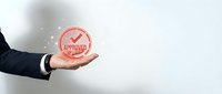 Businessman hand presenting red approved stamp badge with checkmark on light background for quality assurance and certification concepts.