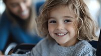 Cheerful young girl with blonde curly hair wearing gray knit sweater smiling brightly at camera, showing genuine happiness and joy in casual indoor setting.
