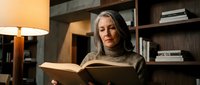 Mature woman with gray hair reading book in cozy home library with warm lighting and wooden shelves for relaxation and learning concepts.