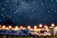 Cozy winter evening with knitted blanket, hot chocolate and marshmallows against dark blue starry sky and warm string lights background.