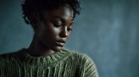Young Black woman with short curly hair wearing olive green ribbed sweater, looking downward against teal studio background. Portrait for fashion, lifestyle, and wellness content.