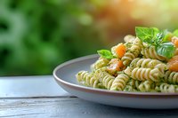 Fusilli pasta with fresh basil pesto sauce and cherry tomatoes served on white plate against blurred garden background, summer Italian cuisine.