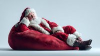 Santa Claus relaxing in red bean bag chair after Christmas delivery, tired holiday character resting with white beard and traditional costume.