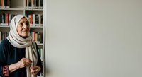 Senior woman wearing beige hijab and dark cardigan stands by bookshelf in library, looking at camera with warm expression. Portrait for education, diversity, and community themes.
