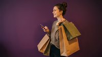 Young woman holding smartphone and shopping bags on purple background, happy consumer with mobile phone after retail therapy session.