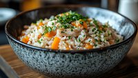 Steamed rice with shrimp and diced carrots garnished with fresh chopped herbs served in textured ceramic bowl on wooden surface, close up food photography.
