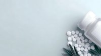 White round pills scattered from medicine bottles with green palm leaves on light gray background, minimalist pharmaceutical composition for healthcare concept.