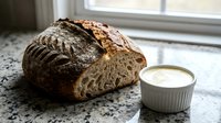 Freshly baked artisan sourdough loaf with crispy crust and scoring marks displayed on granite countertop next to creamy butter in white ceramic dish.