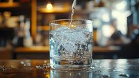 Clear water being poured into glass with ice cubes on bar counter in restaurant setting with warm ambient lighting and bokeh background.