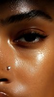 Close-up of brown eye with glowing glitter makeup and dewy skin highlighting cheekbone. Crystal facial piercing adds sparkle to bronzed complexion with glossy finish.