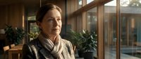 Middle-aged woman with brown hair pulled back, fair complexion, wearing a beige knit sweater and scarf in a modern office or corporate environment with large windows and potted plants, suitable for business, professional, and corporate themes.