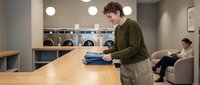 Young Caucasian man folding clean clothes at wooden table in modern laundromat with washing machines and seating area in background.