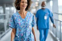 Confident female healthcare worker in floral scrubs walking hospital corridor with male colleague in blue uniform in background.
