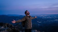 Young man with open arms celebrating freedom on mountain peak at sunset with city lights below during golden hour adventure.