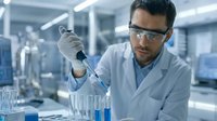 Male scientist in safety glasses using pipette in modern laboratory conducting chemical research experiment with blue liquid samples in test tubes.