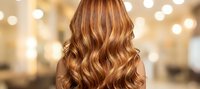 Woman with long wavy auburn hair with golden highlights in professional salon setting with warm lighting for beauty and hair care concepts.