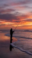 Silhouette of angler fishing at ocean shore during dramatic sunset with colorful sky reflecting on wet sand and gentle waves for outdoor recreation concepts.