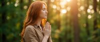 Young Caucasian woman with long red hair in prayer pose outdoors during golden hour sunset, peaceful meditation in nature forest setting for wellness concepts.
