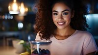 Young African American woman enjoying healthy breakfast with blueberries and yogurt in modern kitchen, smiling at camera with warm lighting and cozy atmosphere.