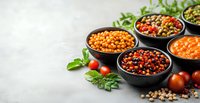 Variety of cooked legumes and beans in black ceramic bowls garnished with fresh basil leaves and cherry tomatoes on light gray concrete background.