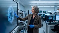 Middle-aged Caucasian female scientist in brown blazer examining blue molecular structure on laboratory monitor wearing protective gloves in modern research facility.