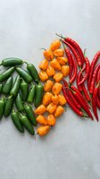 Colorful hot chili peppers arranged on concrete surface featuring green jalapenos, orange habaneros and red cayenne varieties for cooking and spice preparation.