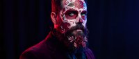 Bearded man with intricate Day of the Dead sugar skull face paint featuring red and white floral designs against dark background for Halloween costume.