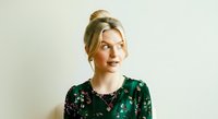 Caucasian woman with blonde hair styled in updo, wearing dark green floral blouse, looking away thoughtfully against neutral background for portrait and lifestyle use.