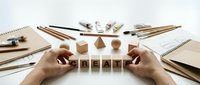 Creative professional arranging wooden letter blocks spelling create surrounded by art supplies, sketchbooks, and design tools on white workspace for artistic projects.