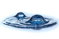 Macro photography of water drops creating crown splash effect with blue tones and ripples on liquid surface, captured at high speed with selective focus.