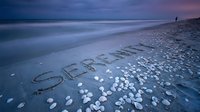 Serenity written in sand on a tranquil beach at dusk, with scattered shells, soft waves, and a lone figure in the distance.