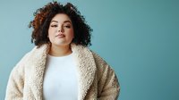 Confident young woman with curly hair wearing cozy sherpa jacket and white top against teal background, showcasing body positivity and modern fashion.