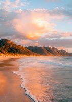Dramatic coastal landscape at golden hour with waves washing onto sandy beach beneath rugged mountains, illuminated by warm sunset light and soft clouds.