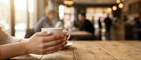 Hands holding white coffee cup on wooden table in cozy cafe with warm lighting and blurred customers in background creating relaxing atmosphere.