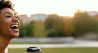 Young woman with curly hair, medium skin tone, laughing outdoors during golden hour with coffee cup and blurred urban park background, expressing joy and relaxation.