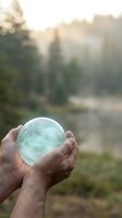 Hands holding glowing crystal ball with soft sunlight rays streaming through blurred forest background creating mystical atmosphere for fortune telling concepts.
