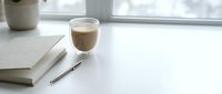 Workspace setup with coffee cup, notebook and pen on white desk near window with plant in background.