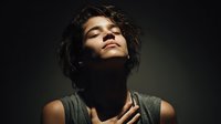 Young woman with curly hair in dramatic lighting, eyes closed in peaceful expression wearing sleeveless top, illuminated from above against dark background.