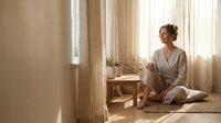 Young woman in comfortable pajamas sitting cross-legged on floor in bright bedroom, enjoying peaceful morning routine with coffee and natural sunlight.