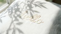 Hello embroidered in cursive on a soft white linen pillow with delicate leaf shadows, creating a warm, cozy home decor atmosphere.