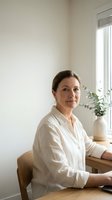 Middle-aged woman with brown hair wearing white linen shirt, seated at wooden desk in bright minimalist interior with potted plant and window light, professional portrait for business and lifestyle use.