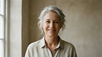 Mature woman with gray wavy hair and fair complexion smiling at camera in bright interior space, suitable for lifestyle, wellness, and professional portraits.