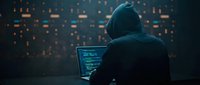 Hooded hacker in dark room working on laptop with glowing code on screen. Cybersecurity threat concept for digital security and data protection awareness.
